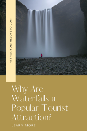 A person in a red jacket standing near the base of a tall, misty waterfall on a cloudy day, with text overlay reading: 'Why Are Waterfalls a Popular Tourist Attraction? Learn more at fortheloveto.com.