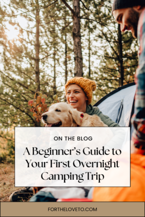 A smiling woman in a cozy knit hat sits by a tent in a forest, holding a golden retriever, while a man in a beanie stands nearby. Overlaid text reads: 'A Beginner’s Guide to Your First Overnight Camping Trip' - fortheloveto.com.