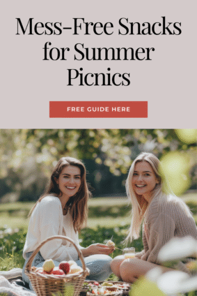 Two women smiling and enjoying a picnic outdoors with a fruit basket and snacks, with text overlay: "Mess-Free Snacks for Summer Picnics – Free Guide Here". Ideal pin for a blog post about must-have picnic snacks and essentials.
