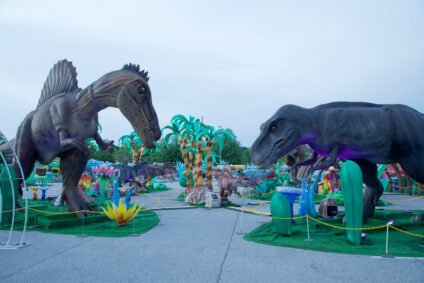 Two towering animatronic dinosaurs—a Spinosaurus and a T-Rex—face each other in a colorful outdoor exhibit area at LuminoCity Dino Safari. The scene is filled with palm trees, plants, and smaller dinosaur sculptures, creating an immersive prehistoric atmosphere.