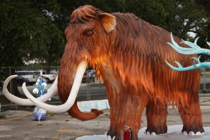 A large illuminated woolly mammoth sculpture with detailed fur texture and long white tusks stands in an outdoor display area. The mammoth is glowing with warm light, surrounded by decorative elements and trees in the background.