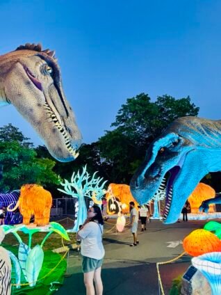 Visitors walking among glowing dinosaur and mammoth light sculptures at LuminoCity Dino Safari Long Island during a nighttime event.