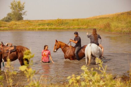 Scene from East of Wall movie showing teens bonding while riding and walking horses in a shallow creek surrounded by fields, showcasing rural life and healing connections.