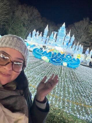Iveth from For The Love To smiling in front of the glowing Lumi Castle at the LuminoCity Holiday Lights Festival in Eisenhower Park, Long Island.