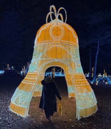 Visitor walking under a giant golden bell light sculpture at LuminoCity Festival, surrounded by sparkling lights and nighttime magic.