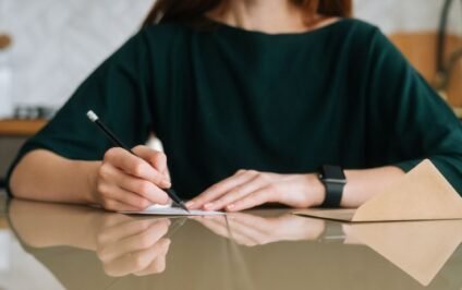 Woman writing a handwritten note at a table, representing how it’s never too late to reach out to someone and reconnect through thoughtful communication.