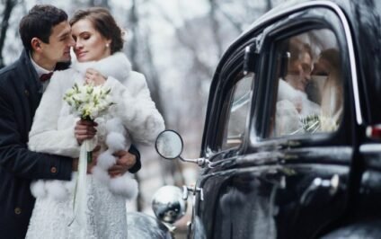 Bride holding a white bouquet beside her groom and a black vintage car, showing how to add vintage charm to a winter wedding